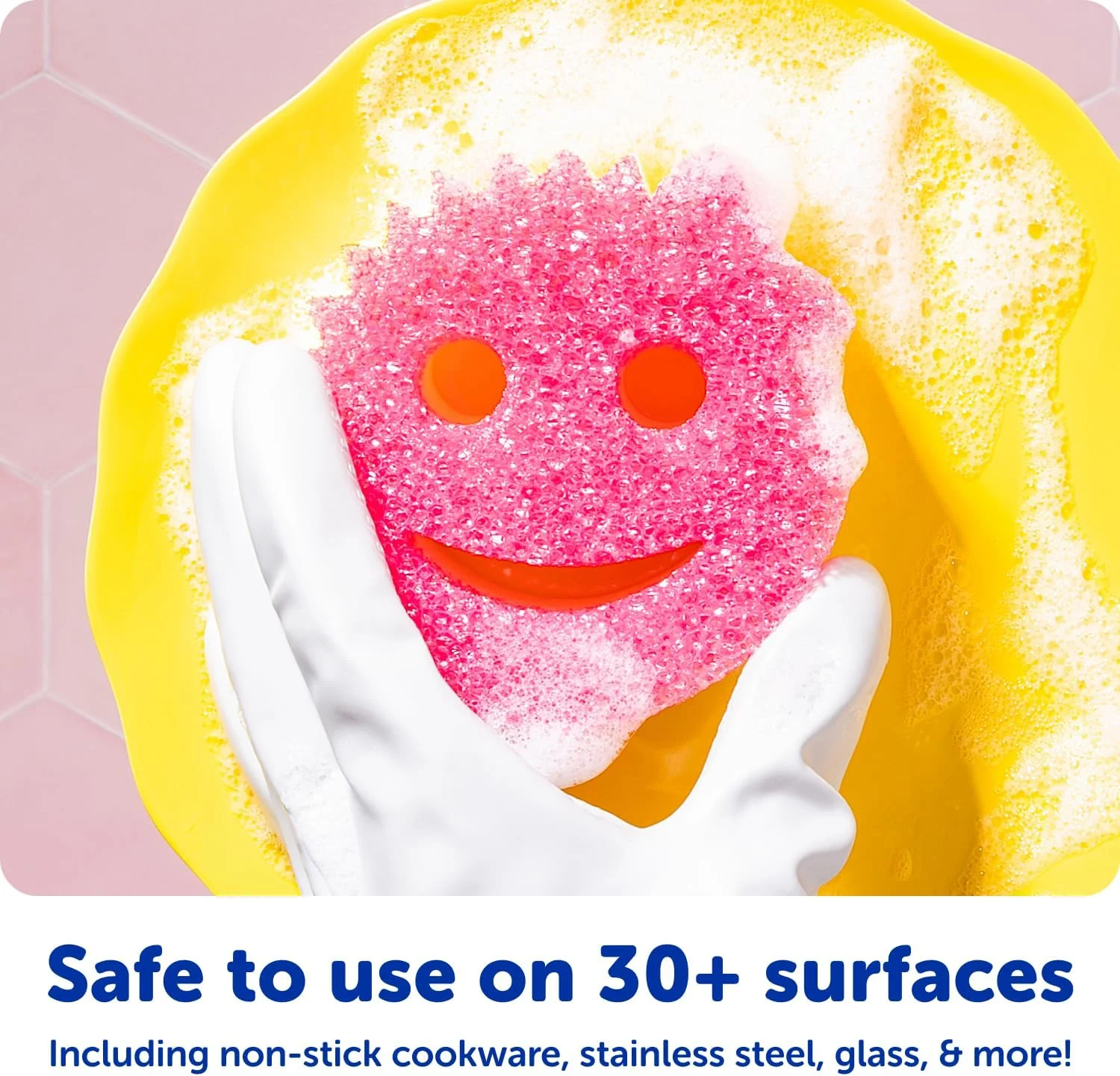 A hand in a white glove holding a soapy pink smiley face sponge over a yellow dish, showing safe use on cookware.